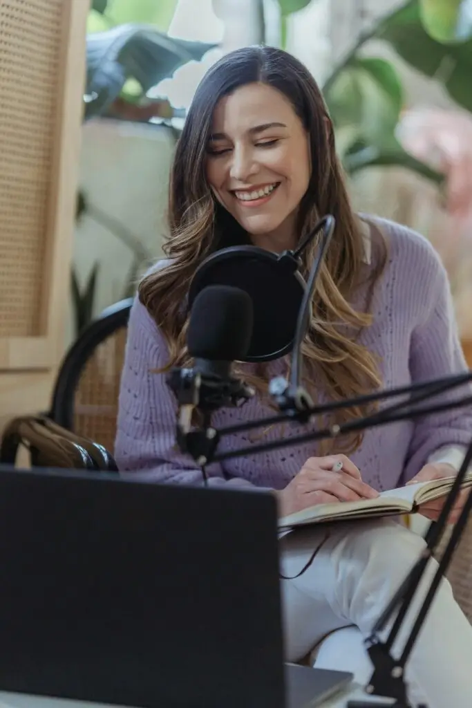 Cheerful woman hosting a podcast from home, using a laptop and microphone setup.